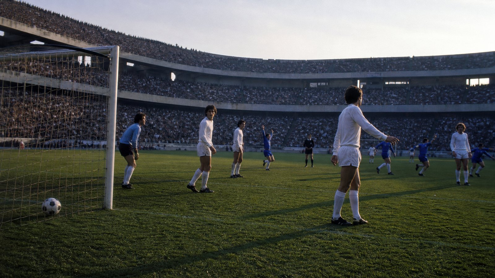 Anonymous footballers in plain blue and white early-1980s kits play under floodlights in a large Spanish stadium